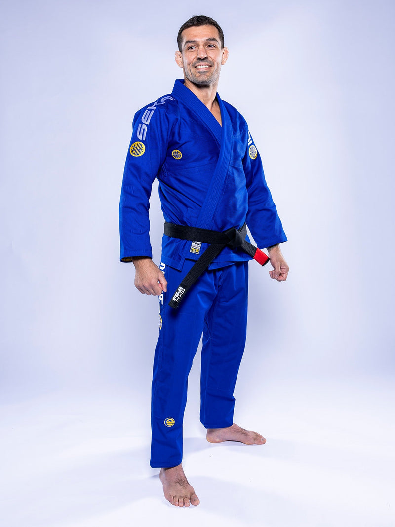 A person stands barefoot on a white background, smiling with arms relaxed at their sides, wearing the SEKAI 3.0 BJJ Gi Blue Yellow and a black belt.