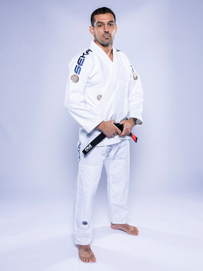 A man in the SEKAI 3.0 BJJ Gi White Navy stands barefoot on a white background, confidently holding a black belt with both hands and looking at the camera.
