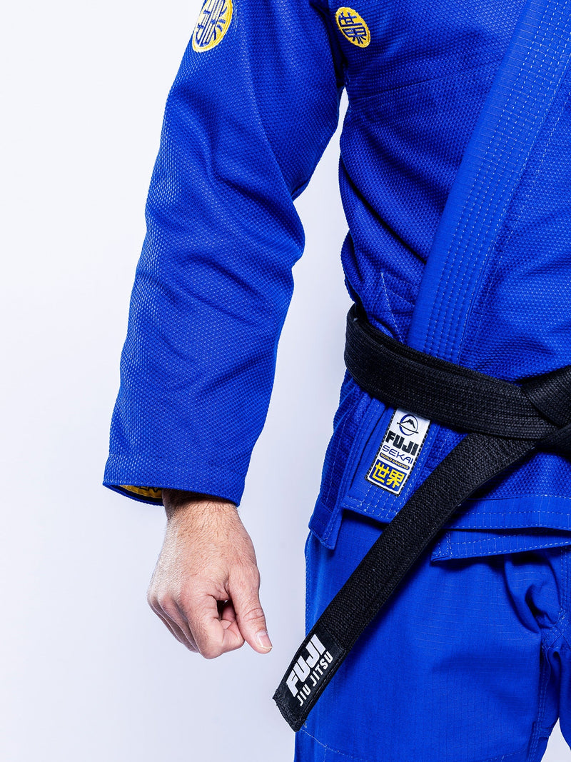 A person wearing the SEKAI 3.0 BJJ Gi Blue Yellow and a black belt, with only their torso and left arm visible against a plain white background.