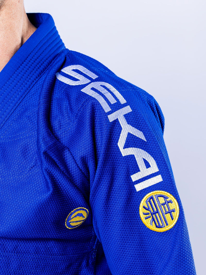 Close-up of a person wearing the SEKAI 3.0 BJJ Gi Blue Yellow, featuring "SEKAI" in white on the shoulder and two yellow circular patches on the upper arm against a plain light background.