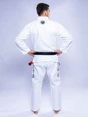 A person in a SEKAI 3.0 BJJ Gi White Navy with a black belt stands barefoot, facing away with hands on hips. The gi features patches and embroidery on the back and legs against a light background.