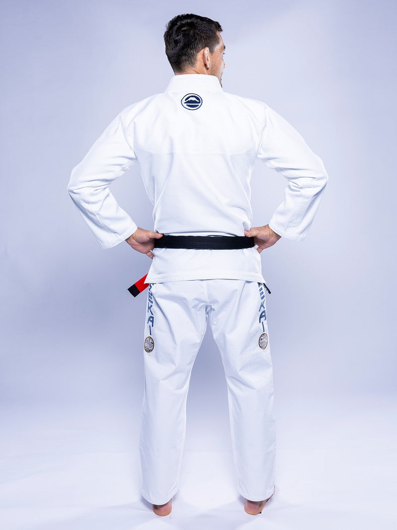 A person in a SEKAI 3.0 BJJ Gi White Navy with a black belt stands barefoot, facing away with hands on hips. The gi features patches and embroidery on the back and legs against a light background.