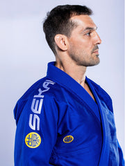 A man wearing the SEKAI 3.0 BJJ Gi Blue Yellow with “SEKAI” on the shoulder and a yellow sleeve patch stands in profile against a light background, facing right.