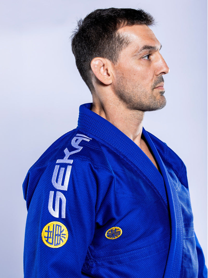 A man wearing the SEKAI 3.0 BJJ Gi Blue Yellow with “SEKAI” on the shoulder and a yellow sleeve patch stands in profile against a light background, facing right.