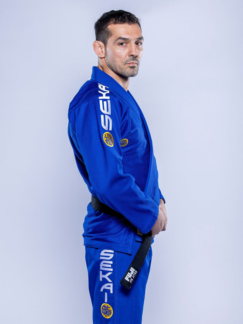 A man wearing the SEKAI 3.0 BJJ Gi Blue Yellow stands in profile, holding his black belt and looking seriously at the camera against a plain light background.