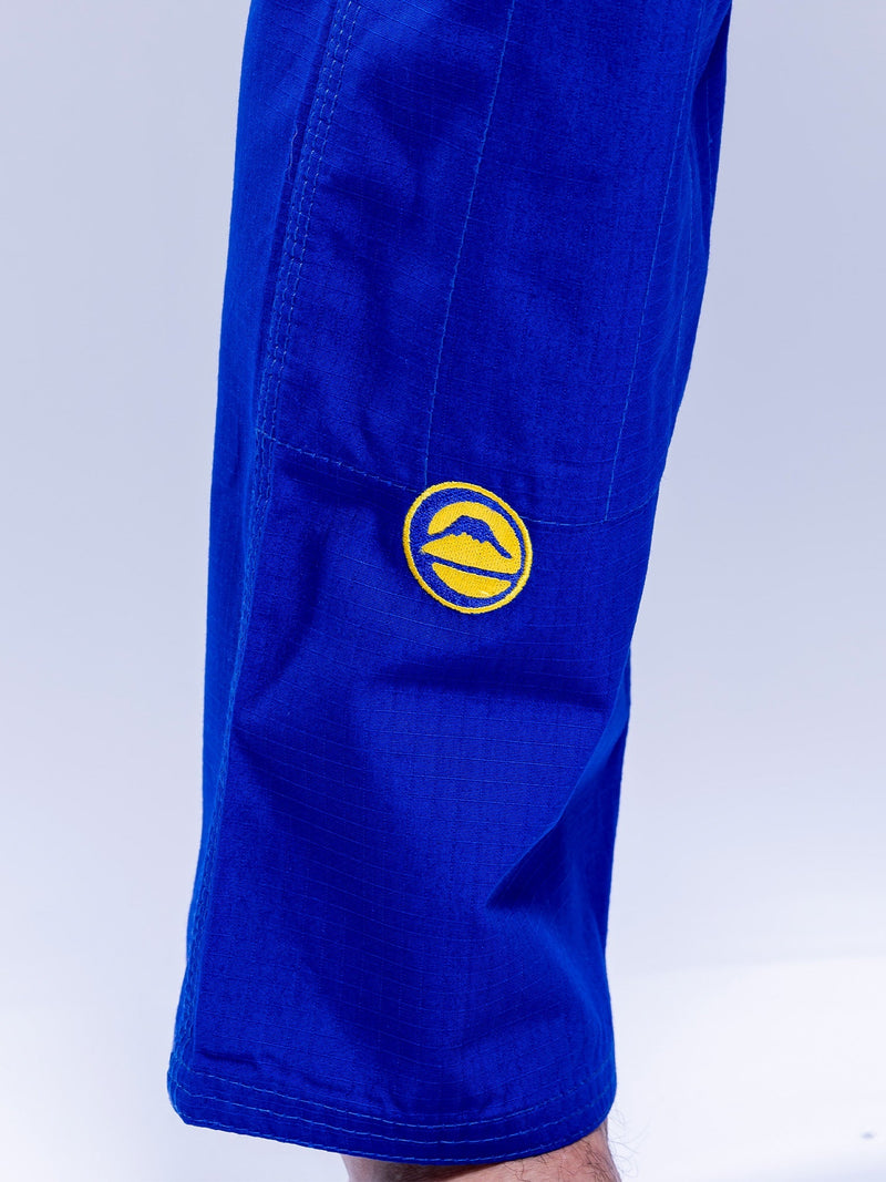 A close-up of someone wearing the SEKAI 3.0 BJJ Gi Blue Yellow, featuring a yellow circular mountain patch on the upper leg. The background is light and uncluttered.