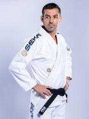 A man stands confidently with hands on hips, wearing the SEKAI 3.0 BJJ Gi White Navy with black belt. The gi features patches and "SEKA" on the sleeve and pants against a light gray background.