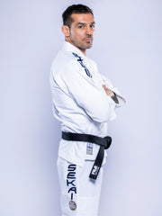 A man wearing the SEKAI 3.0 BJJ Gi White Navy with blue and black lettering and patches, plus a black belt, stands sideways with arms crossed, looking at the camera against a plain light background.