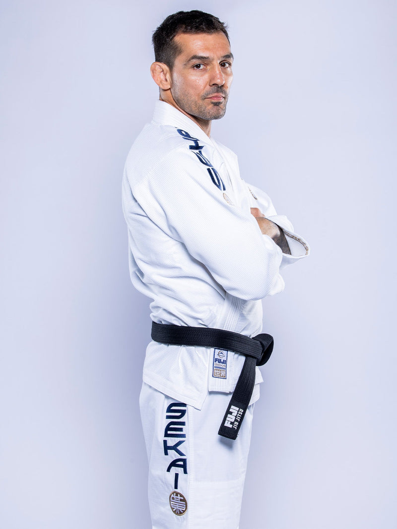 A man wearing the SEKAI 3.0 BJJ Gi White Navy with blue and black lettering and patches, plus a black belt, stands sideways with arms crossed, looking at the camera against a plain light background.