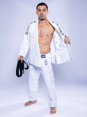 A man stands barefoot on a white floor wearing the SEKAI 3.0 BJJ Gi White Navy with patches, holding a black belt in his left hand and opening his jacket with his right to reveal his chest.