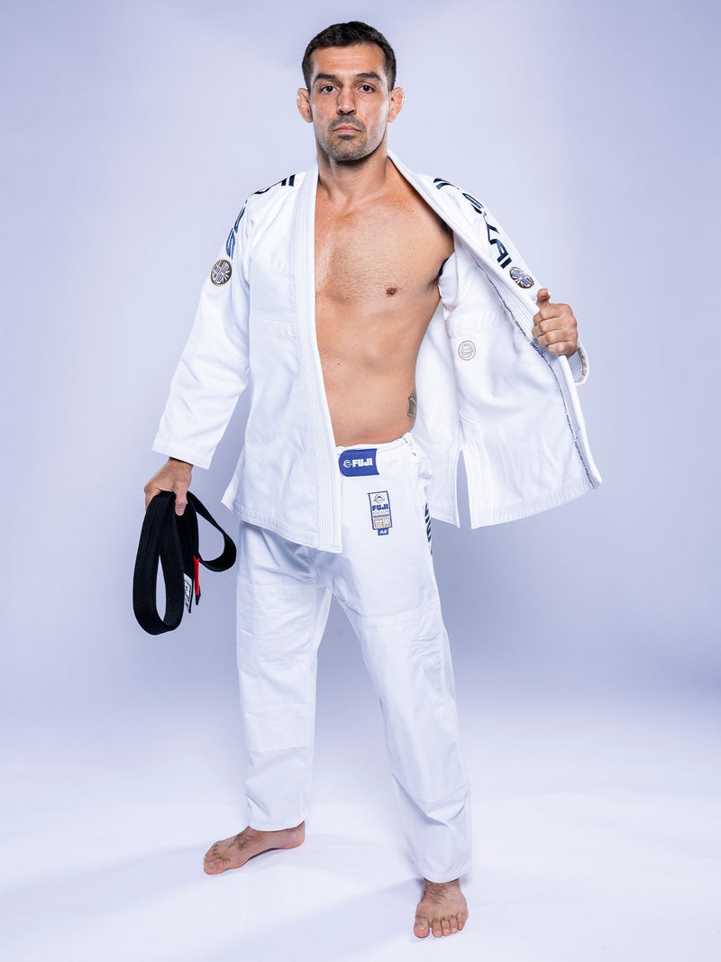 A man stands barefoot on a white floor wearing the SEKAI 3.0 BJJ Gi White Navy with patches, holding a black belt in his left hand and opening his jacket with his right to reveal his chest.