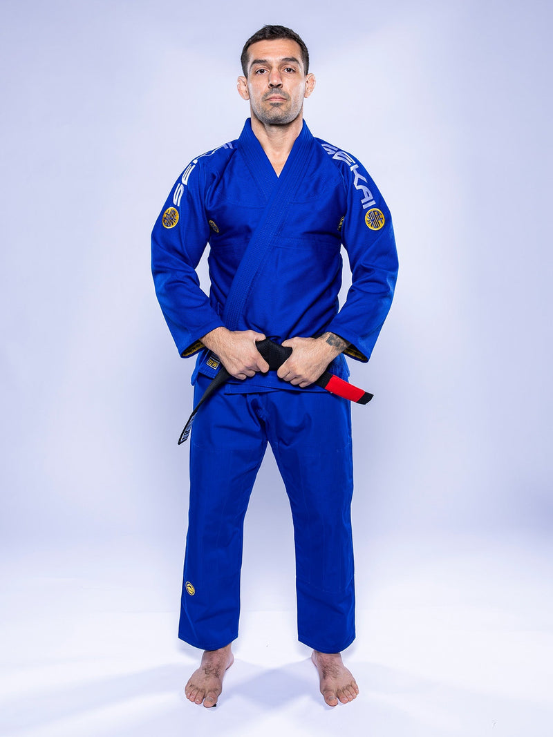 A man wearing the SEKAI 3.0 BJJ Gi Blue Yellow stands barefoot on a white background, holding a black belt with red bar at his waist and looking straight ahead with a neutral expression.