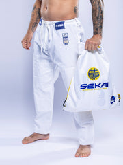 A person in a SEKAI 3.0 BJJ Gi White Navy holds a matching white drawstring bag labeled SEKAI WORLD RANKING with blue and yellow logos, standing barefoot against a light background.