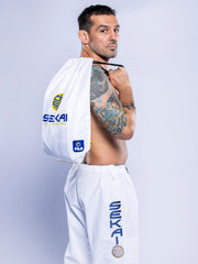 A man with short dark hair and a tattooed right arm poses in SEKAI 3.0 BJJ Gi White Navy pants, holding a white SEKAi drawstring bag over his shoulder against a plain light background.