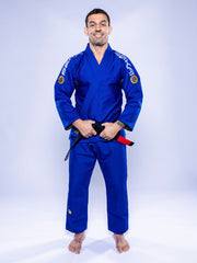 A man stands barefoot on a white background, wearing the SEKAI 3.0 BJJ Gi Blue Yellow and holding a black belt with a red bar while smiling at the camera.