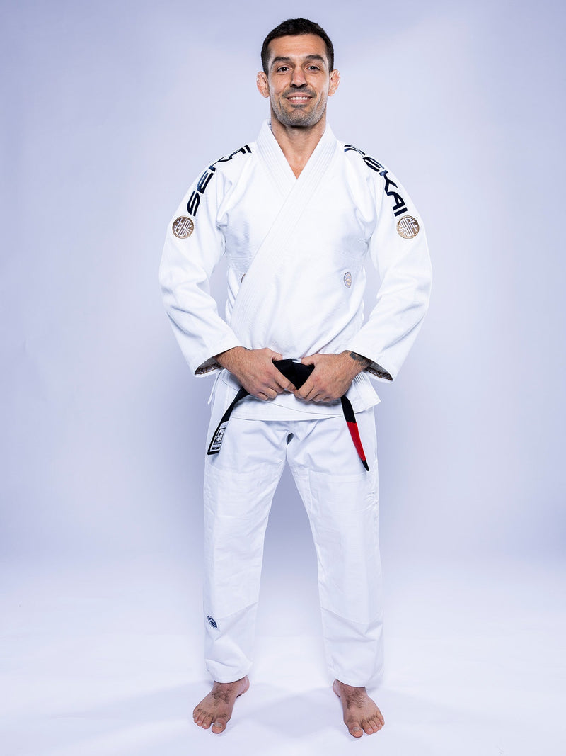A man wearing the SEKAI 3.0 BJJ Gi White Navy with a black belt stands barefoot on a white background, smiling and facing forward with his hands resting on his belt.