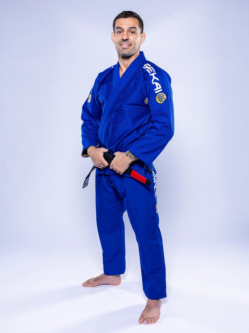 A man stands barefoot on a white background, smiling while wearing the SEKAI 3.0 BJJ Gi Blue Yellow with patches and holding his black belt tied around his waist.