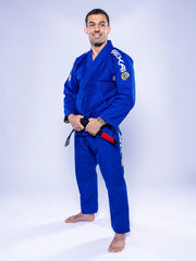 A man wearing the SEKAI 3.0 BJJ Gi Blue Yellow with a black belt stands barefoot on a white background, smiling and facing the camera.