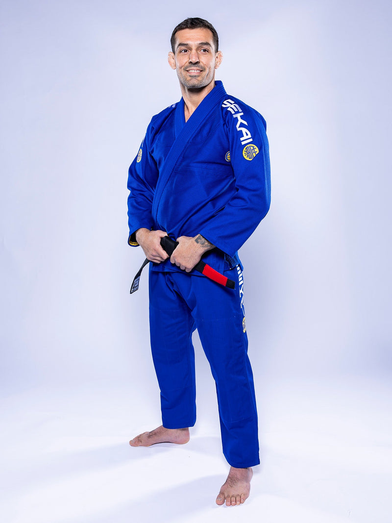 A man wearing the SEKAI 3.0 BJJ Gi Blue Yellow with a black belt stands barefoot on a white background, smiling and facing the camera.
