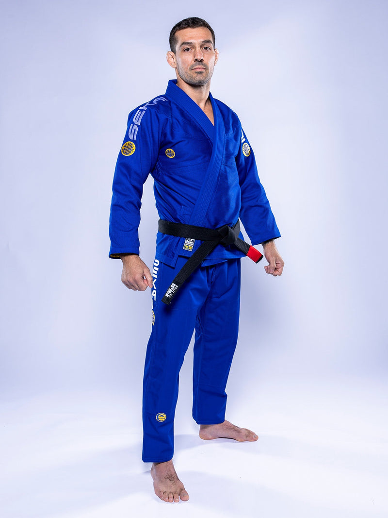 A man wearing the SEKAI 3.0 BJJ Gi Blue Yellow with a black belt stands barefoot on a white background, facing forward with a serious expression.