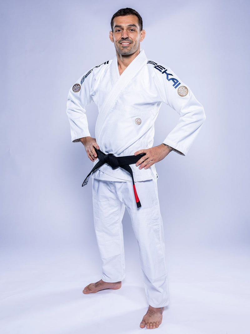 A person wearing the SEKAI 3.0 BJJ Gi White Navy and a black belt stands barefoot on a white background, smiling with hands on hips.