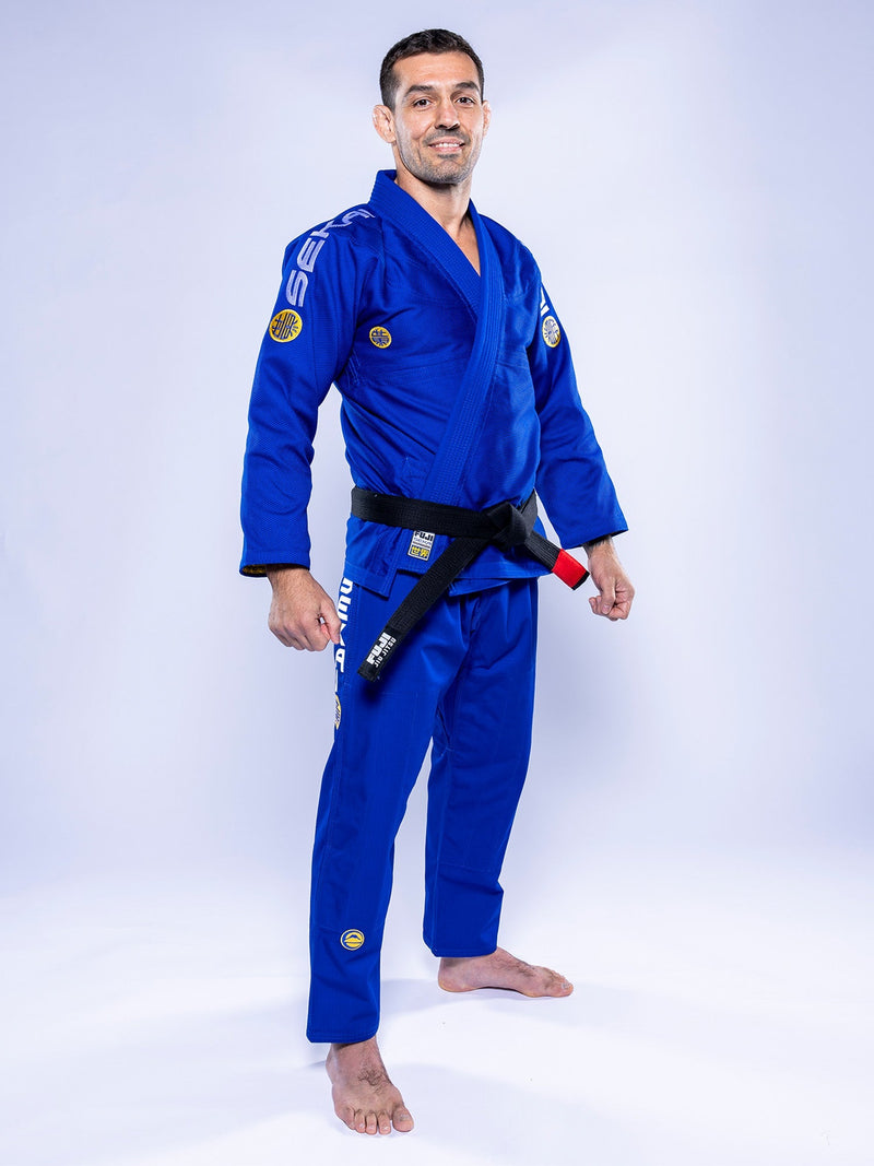 A man stands barefoot on a white background, wearing the SEKAI 3.0 BJJ Gi Blue Yellow with patches and a black belt, smiling and facing forward.